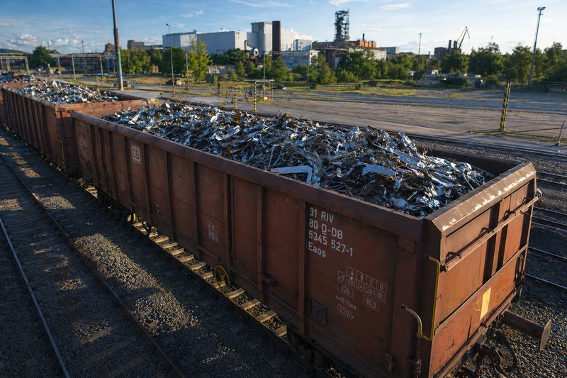 An open freight car loaded with scrap metal stands on the tracks. Next to it is a large pile of scrap metal and seven rolls of steel strip.