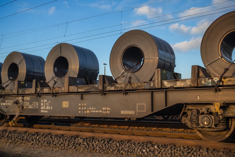 A flat car loaded with coils, i.e., flat-rolled, strip-shaped steel sheet on rolls, is standing on a track. The coils are unloaded using a crane.